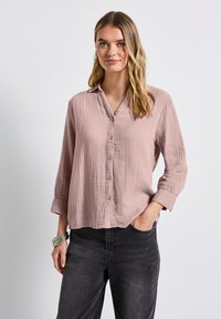 Frau in einer lockeren, staubigen rosa Knopfleiste-Bluse mit 3/4-Ärmeln, kombiniert mit schwarzen Jeans, silbernen Armbändern und Creolen.