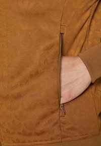 Veste zippée marron avec un motif de feuilles subtil, dotée de poignets en côtes et d'une poche latérale sur le côté gauche, fabriquée dans un tissu texturé.