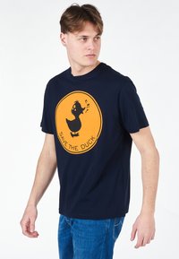 T-shirt di cotone blu navy con una grafica circolare arancione raffigurante un'anatra e il testo "SALVA L'ANATRA". Maniche corte, vestibilità rilassata.