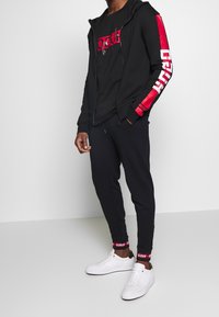 Black zip-up hoodie with red stripes and logos, paired with a black T-shirt and joggers, complemented by white sneakers.