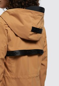 Giacca beige impermeabile con cappuccio, cinturini neri con branding e texture liscia. Ideale per attività all'aperto o per un look casual.