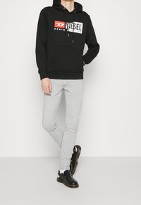 Sudadera negra con bolsillo frontal, con un logo blanco y detalles rojos. Combinada con jeans ajustados gris claro y zapatos negros.