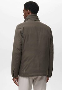 Man die met zijn rug naar ons toe staat, gekleed in een donkergroene jas met hoge kraag en lichte beige broek, tegen een eenvoudige witte achtergrond.