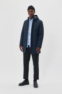 Parka impermeable de color azul marino con capucha, cierre frontal y botones a presión, llevada sobre una camisa azul clara y pantalones negros. Zapatos negros completan el atuendo.