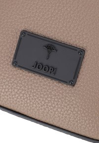 Superficie in pelle beige testurizzata con un'etichetta logo rettangolare nera che presenta la parola "JOOP!" e un emblema decorativo.