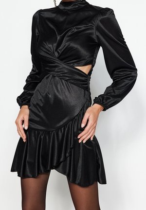 Vrouw draagt een zwart satijnen mini-jurkje met lange mouwen, een twistdetail en een zijopengewerkt, gecombineerd met doorschijnende zwarte panty's.