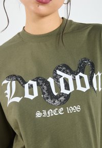 Oliivroheline T-särk, millel on valge gooti stiilis kiri "London" ja musta miku graafika, all aastaarv "1998". Puuvillane kangas, mugav lõige.
