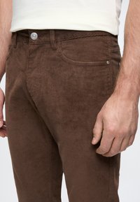 Καφέ καμπάνες από κοτλέ με ριχτό υφή, διαθέτουν μπροστινές τσέπες και μεταλλικό κούμπωμα. Κοντινό πλάνο που δείχνει τη μέση και τη θέση του χεριού.