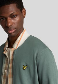 Veste bomber verte à zip, avec un fini texturé, arborant un logo brodé jaune. Portée par-dessus une chemise à carreaux avec des accents orange.