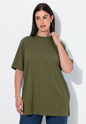 Frau mit langen dunklen Haaren, die ein lockeres olivgrünes T-Shirt und blaue Jeans trägt, schaut seitlich mit einer Hand nahe am Hals.