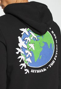 Sudadera negra con un gráfico de la Tierra en azul y verde, rodeada de pájaros blancos, y el texto "REPIENSA - EL FUTURO ES AHORA."