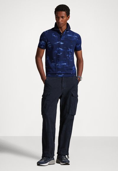 Tamsiai mėlynas polo marškinėlis su mėlynu kalnų raštu, trumpomis rankovėmis ir apykakle; derinamas su tamsiomis krovininėmis kelnėmis ir sportiniais sportbačiais.