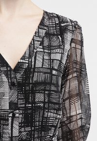 Blusa a pattern bianco e nero con scollo a V e bottoni, dotata di maniche lunghe trasparenti e disegni astratti texture in rete.