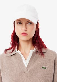 Weiße Baumwollmütze mit gebogenem Schirm, getragen mit einem beigen Pullover mit V-Ausschnitt und grünem Krokodil-Logo auf der Brust. Rotes Haar sichtbar.