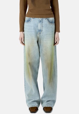 Kvinde iført lyseblå jeans med vide ben og gulligt falmning foran, brun bluse med lange ærmer og brune sko med firkantet tå.