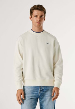 Junger Mann, der ein weißes Sweatshirt mit Rundhalsausschnitt und kleinem Logo auf der Brust sowie hellblaue Jeans trägt, vor einem schlichten Hintergrund stehend.