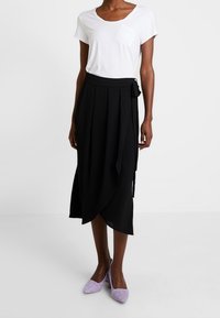 Svart wrapkjol med veck och en knuten detalj, parat med en vit kortärmad skjorta och lila mockaskor med klack. Slät tygfinish.