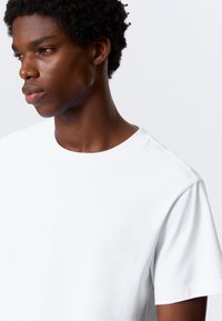 Balta kokvilnas T-krekls ar klasisku apaļu kaklu un īsām piedurknēm, ar gludu tekstūru un minimālistisku dizainu.