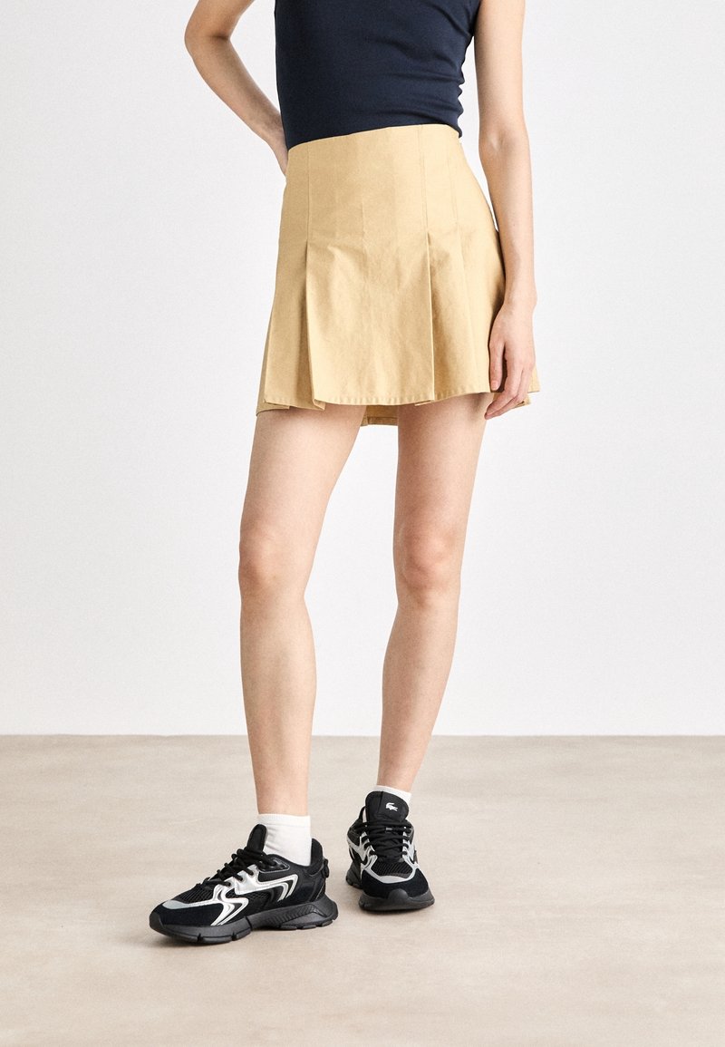 Gonna mini plissettata beige realizzata in tessuto con una texture liscia. Abbinata a sneakers nere con dettagli bianchi e calzini alla caviglia.