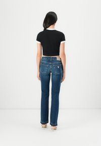 Ženska stoji obrnjena stran od kamere, oblečena v črno skrajšano majico, ravne modre kavbojke in bež sandale na peto, na bež ozadju.