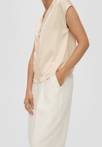 Béžová bezrukávová blůza s výstřihem do V, hladkým povrchem a drapovaným designem, spárovaná s bílými kalhotami s bočními kapsami.