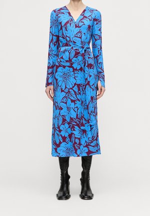 Kobieta ubrana w sukienkę kopertową z długim rękawem, ozdobioną dużym niebieskim kwiatowym wzorem na fioletowym tle, w połączeniu z czarnymi kozakami do kolan.