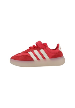 Zapatilla deportiva roja con rayas blancas, detalles en ante, tira de velcro, cordones y suela gruesa translúcida sobre fondo blanco.