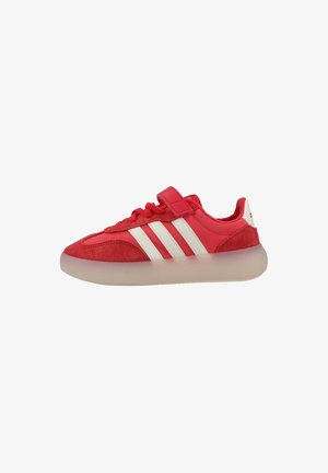 Zapatilla deportiva roja con rayas blancas, detalles en ante, tira de velcro, cordones y suela gruesa translúcida sobre fondo blanco.
