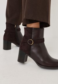 Bottes à cheville en cuir brun avec une sangle en suédine et un accent en métal rond. Design à talon bloc, légèrement texturé avec une fermeture éclair sur le côté.