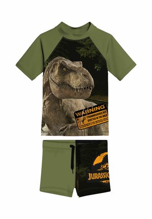 Grüne und schwarze Kinderkleidung mit einer T. Rex-Grafik und dem Schild „Warnung Tyrannosaurus Rex gesichtet“ auf dem Shirt, schwarze Shorts mit Jurassic Park-Logo.