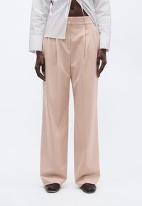 Széles szárú nadrág lágy barackszínű anyagból, magas derékkal, elöl hajtásokkal és sima textúrával. Sötét lábbelivel párosítva.