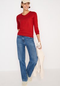 Rotes, figurbetontes Langarmoberteil mit V-Ausschnitt, kombiniert mit blauen, lässig geschnittenen Jeans. Das Model hält einen cremefarbenen, strukturierten Pullover und trägt helle Sneaker.