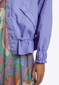 Světle fialová bunda se zipem vpředu, elastickým pasem a manžetami, obsahující cvokovou kapsu a lehkou, texturovanou látku.