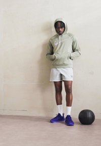 Kapucňová olivová mikina s bílým logem, světle šedé kraťasy a fialové sportovní boty. Černý míč na podlaze. Neutrální pozadí.
