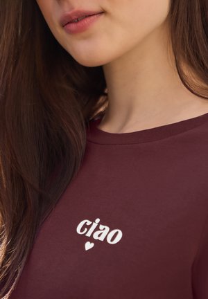 Kobieta z długimi brązowymi włosami, ubrana w bordową koszulkę z napisem „ciao” i małym serduszkiem poniżej.