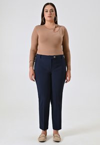 Top beige a maniche lunghe con un dettaglio a fiocco sulla spalla, abbinato a pantaloni tailored blu navy e mocassini beige. Texture liscia, design aderente.