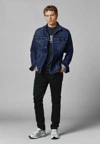 Giubbotto di jeans blu scuro, t-shirt nera con testo, pantaloni neri e sneakers grigie con dettagli bianchi. Modello in piedi con le mani nelle tasche.