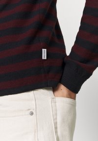 Personne portant un pull rayé noir et rouge foncé avec une étiquette blanche "JACK & JONES", main dans la poche d'un pantalon blanc cassé.
