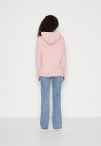 Žena s kudrnatými vlasy, oblečená v růžové mikině a světle modrých džínách, stojící čelem k jednoduché bílé zdi.