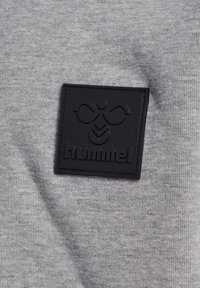 Grått tyg med en subtil textur och en svart gummiloggopatch med ordet "hummel" och en abstrakt design.