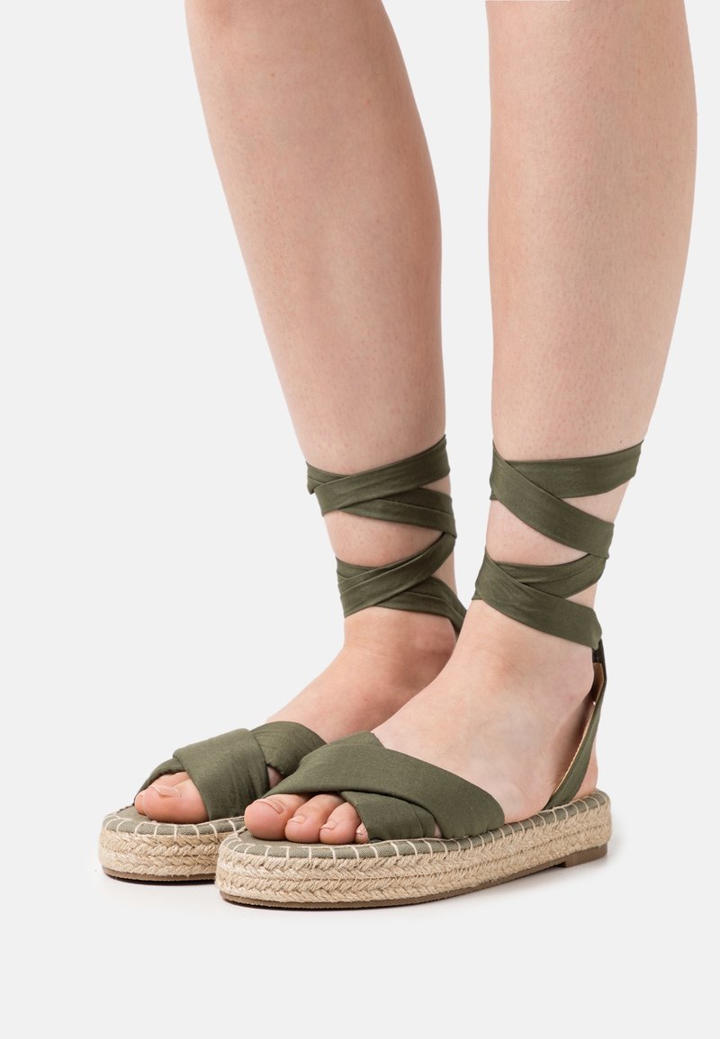 Pieds portant des sandales espadrilles vert olive avec des brides en tissu enroulées autour des chevilles et des semelles en jute tressée sur un fond clair.