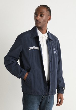 Navy blue coach jacket with a button front, embroidered "Dallas Cowboys" logo, elastic cuffs, and side pockets. Worn over a white shirt.