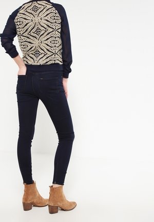 Persoon draagt donkere skinny jeans, beige suède enkellaarzen en een marineblauw jasje met een ingewikkeld crèmekleurig bloemenpatroon op de rug.