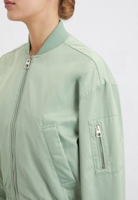 Giacca bomber verde con una superficie liscia, colletto a coste e tasche laterali. Presenta una chiusura lampo frontale e una tasca con zip aggiuntiva sulla manica.