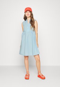 Vestido sem mangas de um azul claro, com fecho de botão e cintura franzida. Combinado com sandálias vermelhas brilhantes de tiras e um boné a condizer. Textura suave.