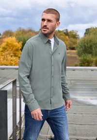 Långärmad, knappad skjorta i mjuk grön tyg, med krage och en bröstlogotyp. Bärs tillsammans med blå jeans utomhus.