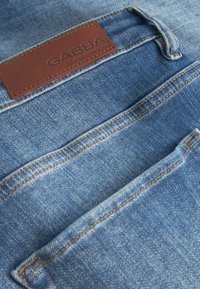 Denimsko blago v svetlo modri barvi z kontrastnimi oranžnimi šivi. Ima rjavo usnje oznako s tekstom "GABBA". Vključuje dva zadnja žepa.