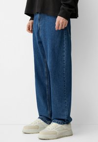 Pánske modré denimové džínsy s voľným strihom, so strižným strihom rovného nohavice, stredným pásom a bežnými detailmi vrecka. Skombinované s bielymi teniskami.