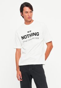 Mand iført en hvid T-shirt med sort tekst, der står "AIN’T GOT NOTHING IT’S GETTING BETTER," sammen med sorte bukser mod en hvid baggrund.