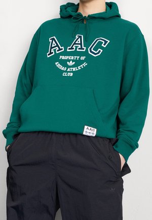 Sweat à capuche vert avec une poche kangourou à l'avant, arborant le texte et le logo "AAC" en blanc. Assorti à un pantalon noir ample, présentant un léger éclat.